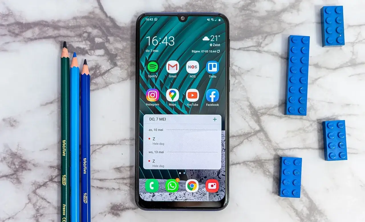 Smartphone displaying various apps on a marble surface with colored pencils and building blocks nearby.