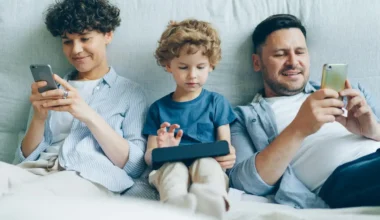 A family of three sitting on a couch, each using a mobile device. The mother and father are on smartphones, while the child uses a tablet.
