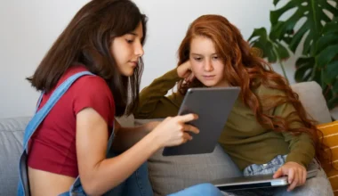 Two young girls sitting on a couch, one holding a tablet while the other looks on attentively.