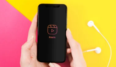 A smartphone held in hands showing the Reels interface on a colorful background with earbuds nearby.